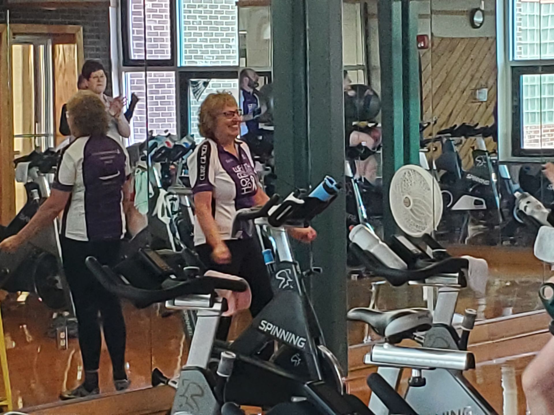 Woman smiles while cycling in a fitness studio with mirrors. Other people and equipment are visible.