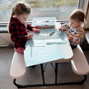 Two children drawing at a small table indoors; one in red plaid shirt, one in floral.