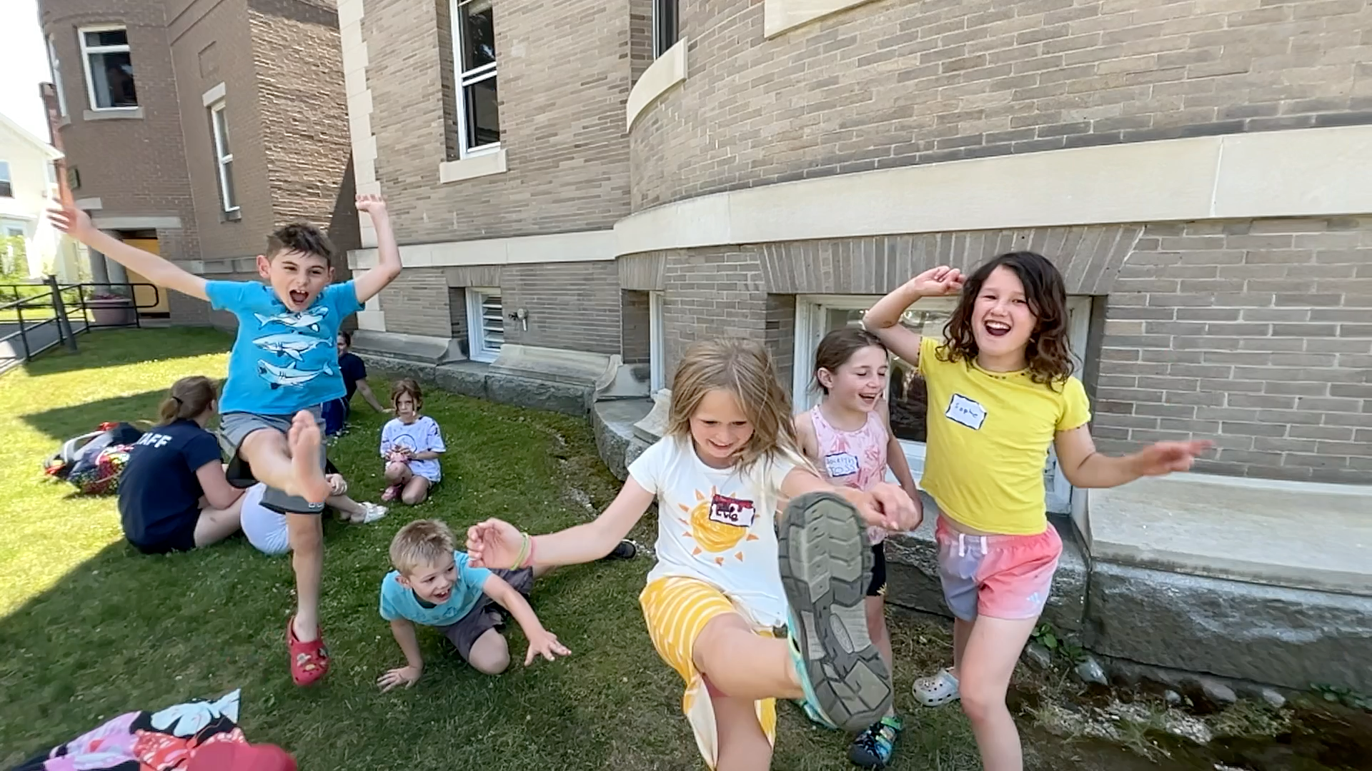 Children playing exuberantly on grass near a brick building. One kicks a dirty shoe at the camera.
