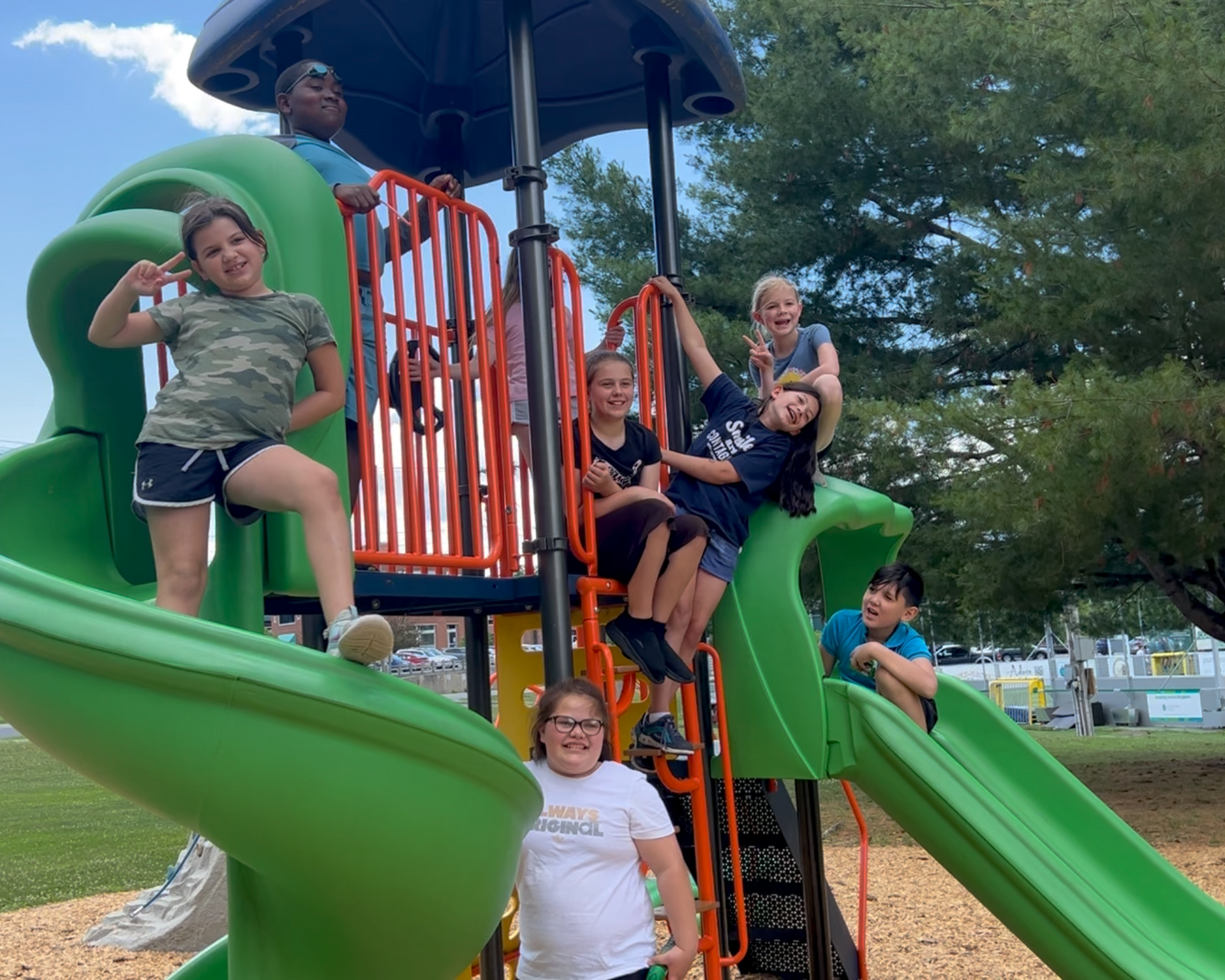 Children on a green playground slide, smiling and making playful gestures. Blue sky in the background.