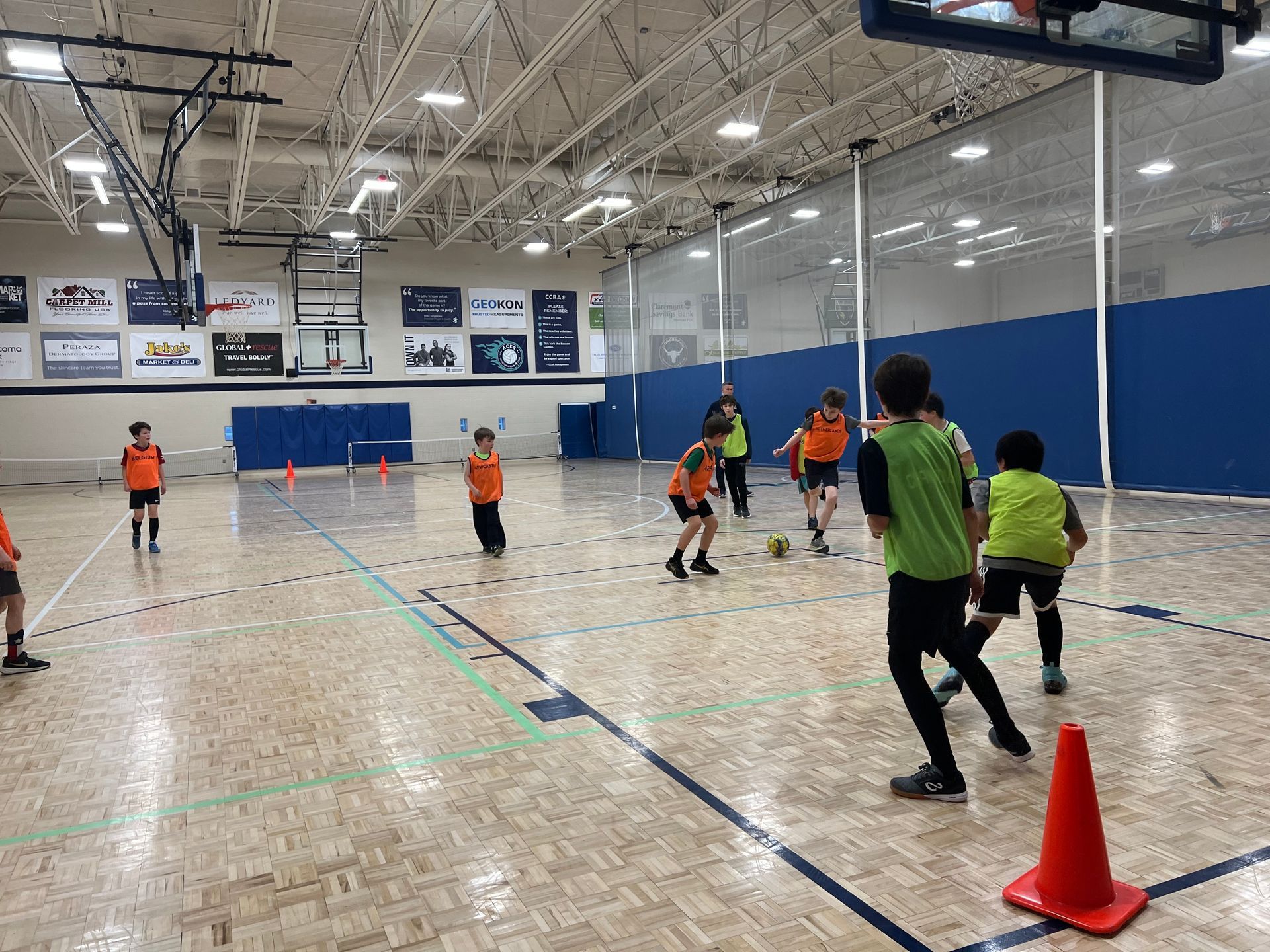 Children playing soccer in a gym, wearing orange and green vests.