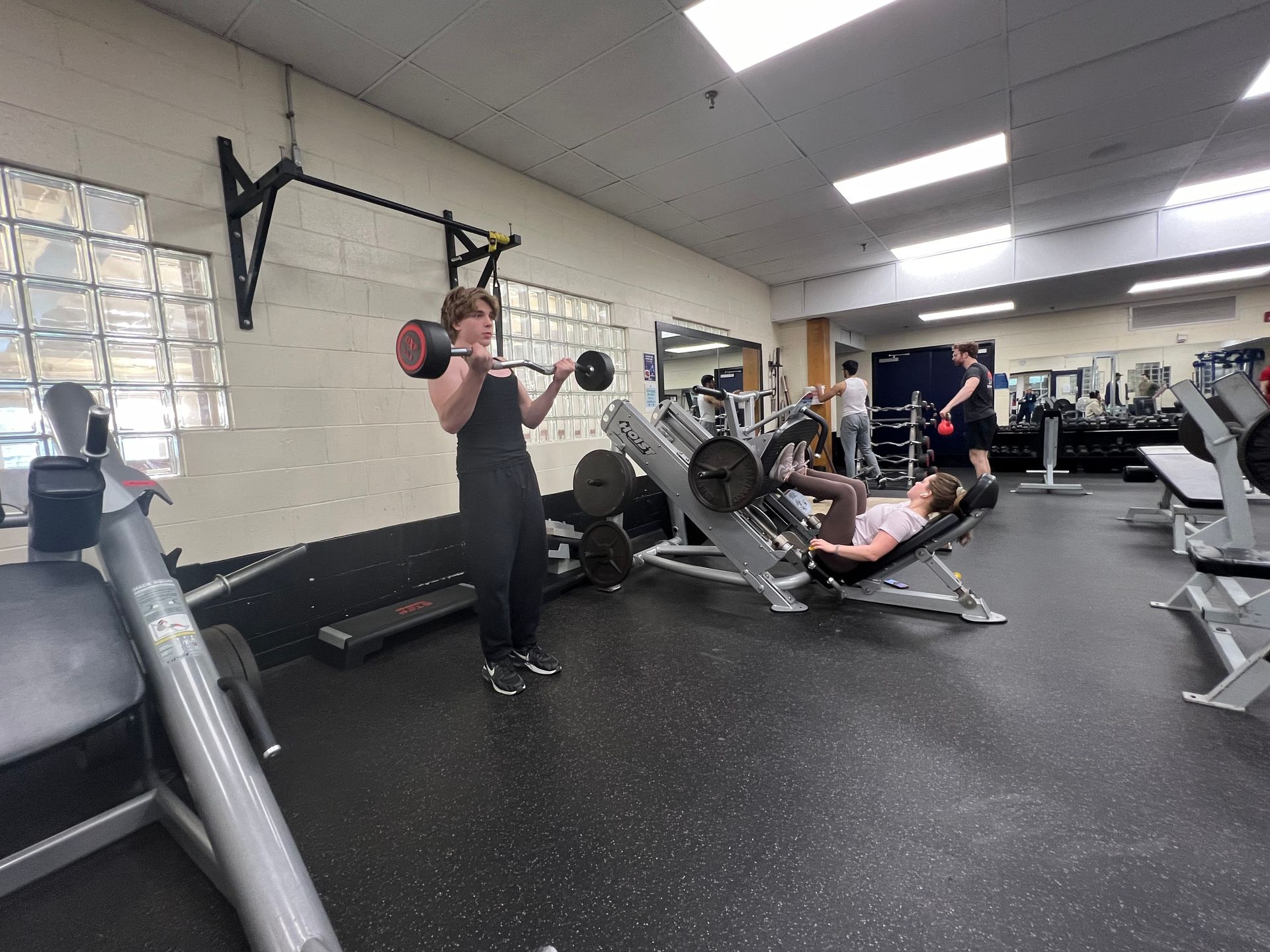 Person lifting barbell in gym, another using leg press machine. Other equipment visible.