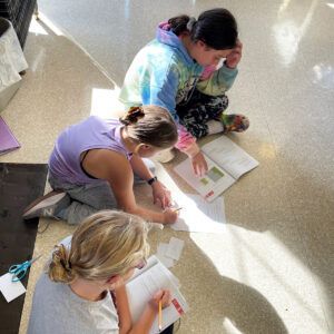 Three people sit on the floor, studying books in a sunlit area, writing and looking.