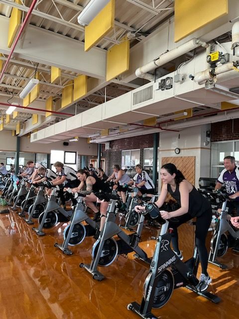 People cycling in a gym. Many exercise bikes are in a large room with yellow ceiling panels and windows.