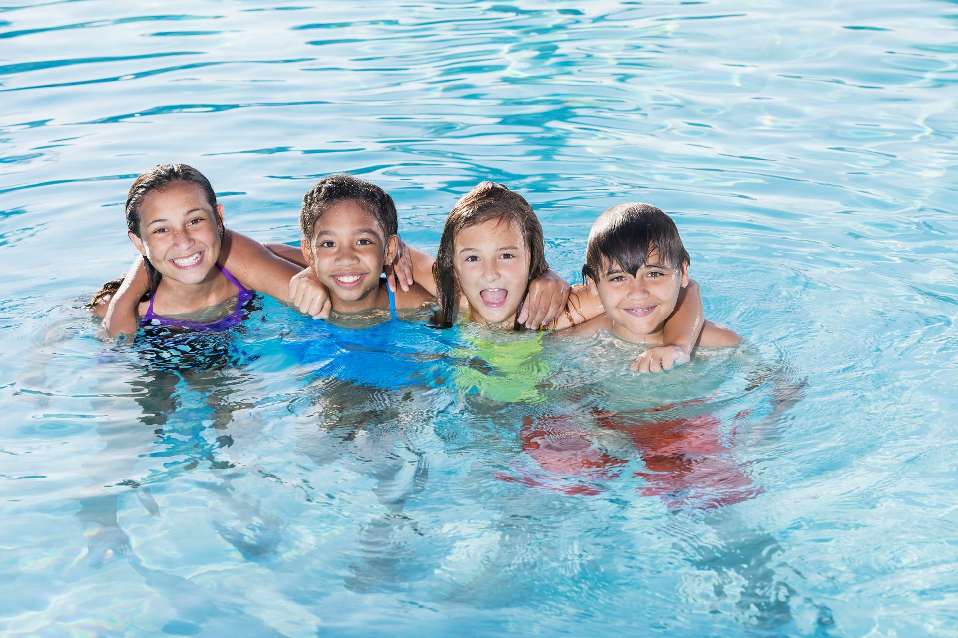 Four children smiling and playing in a blue swimming pool, arms around each other.
