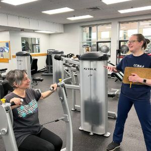 Woman using a weight machine while another person with a clipboard observes in a gym.