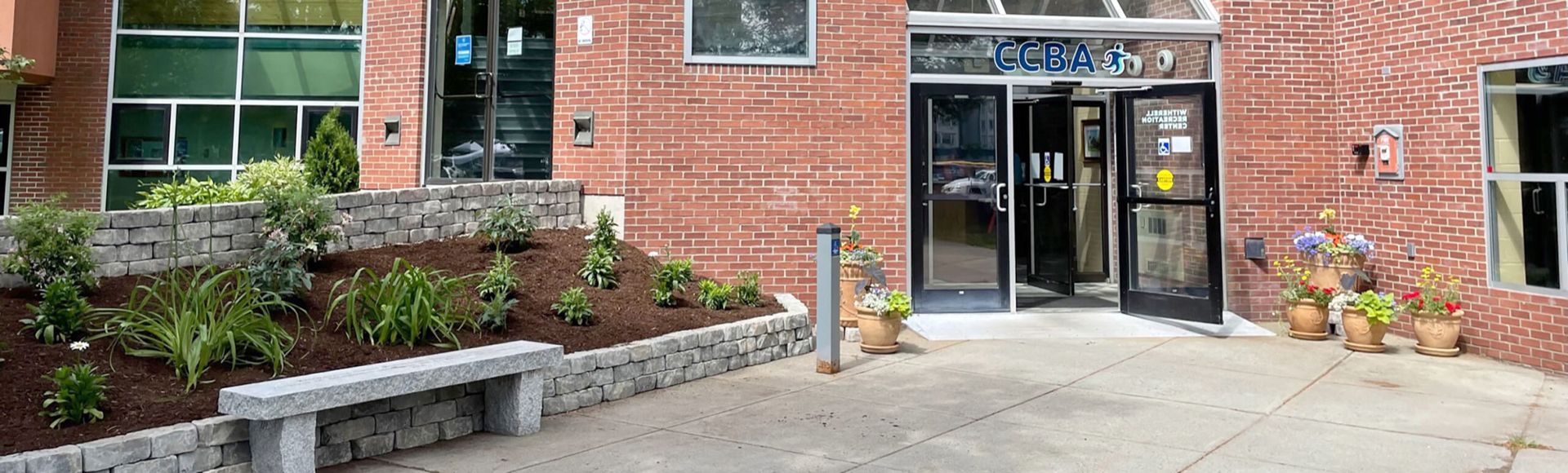 Building entrance with red brick facade, glass doors, and landscaping with a stone bench and planters.