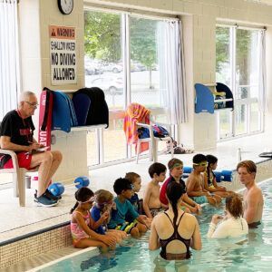 Children in swimsuits sit at pool edge listening to instructors.