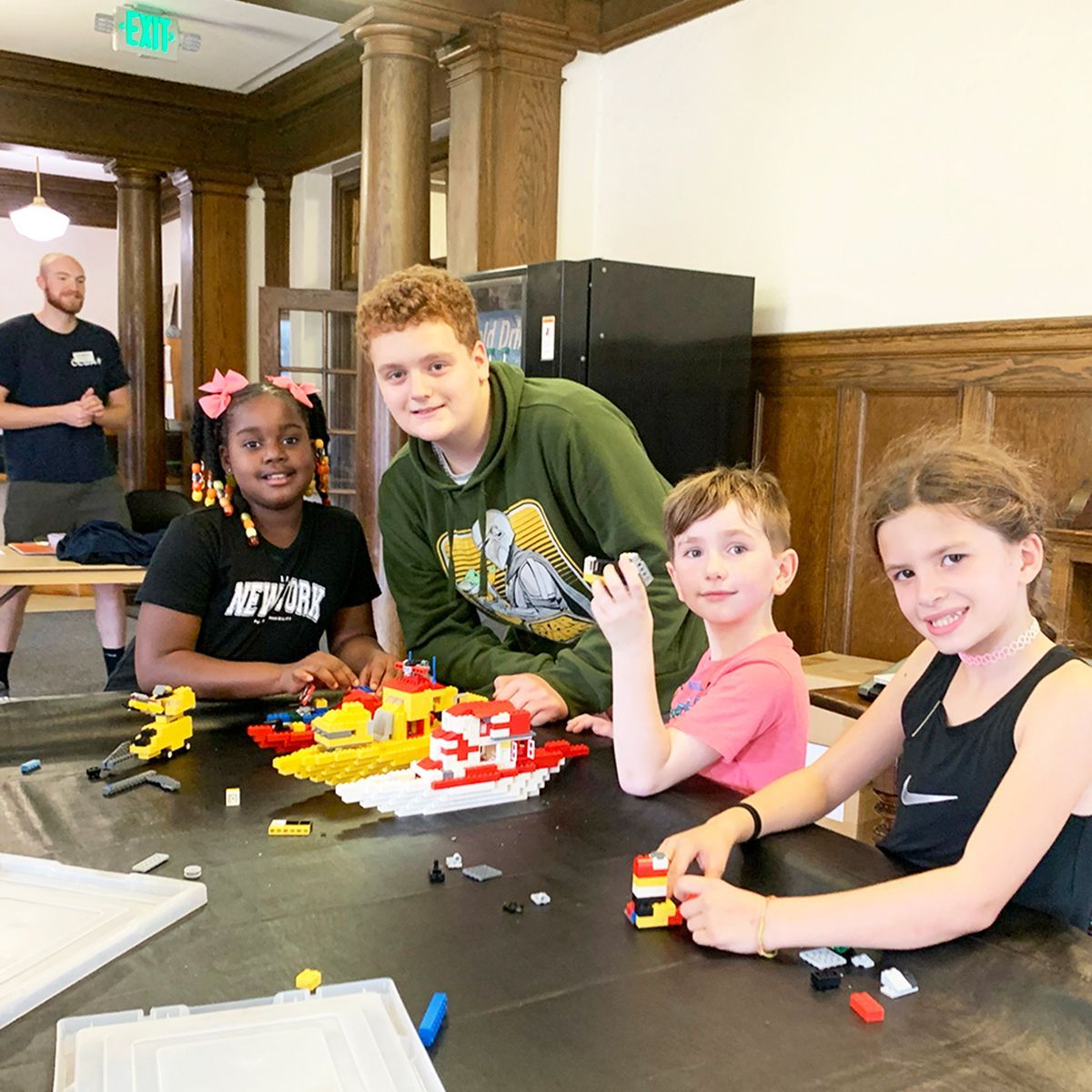Children and a teen building with Legos at a table, with an adult watching.