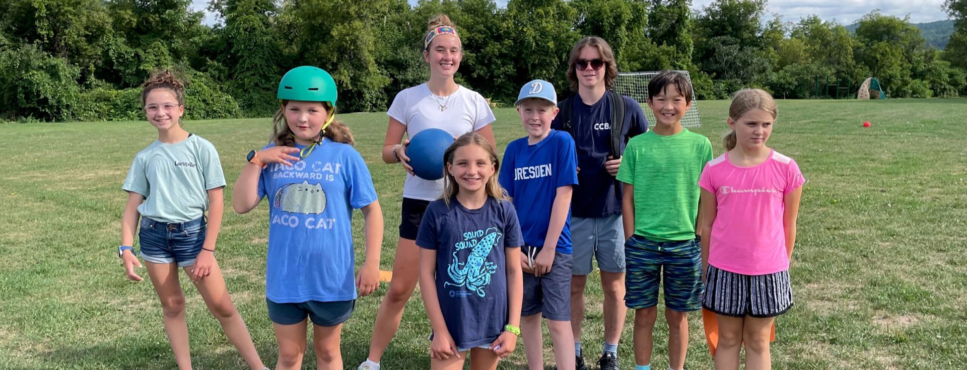 A group of children and an adult pose on a grassy field, some holding balls, trees in the background.