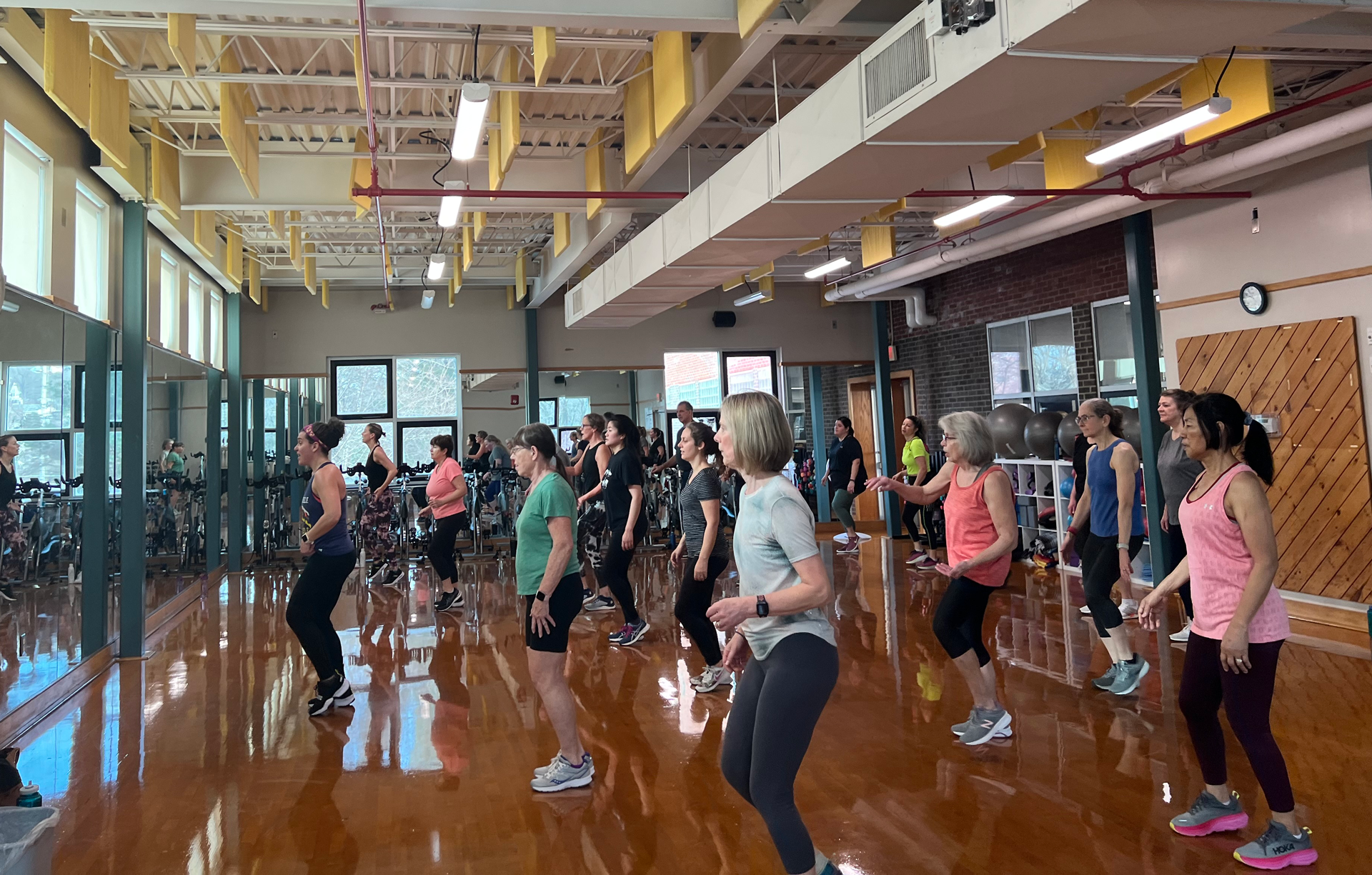 People in a gym exercise class stand on mats, lifting small dumbbells straight overhead with focused expressions.