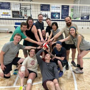 Volleyball team huddles together, arms reaching for a ball. People are smiling inside a gym near a net.