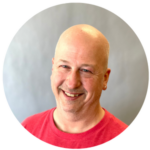 Bald man smiling, wearing a red shirt, set against a neutral gray background.