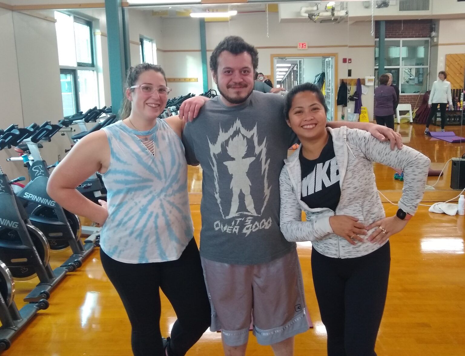Three people smiling, posing in a gym. Woman in tie-dye tank top, man in gray shirt, woman in Nike sweatshirt. Spin bikes in background.