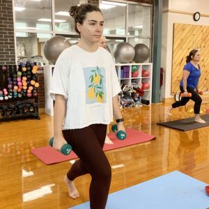 Woman in a gym doing lunges with dumbbells, wearing a t-shirt and leggings. Another woman also exercises with weights.
