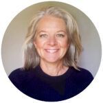 Woman with gray hair smiles, wearing a dark blue sweater, against a cream-colored background.