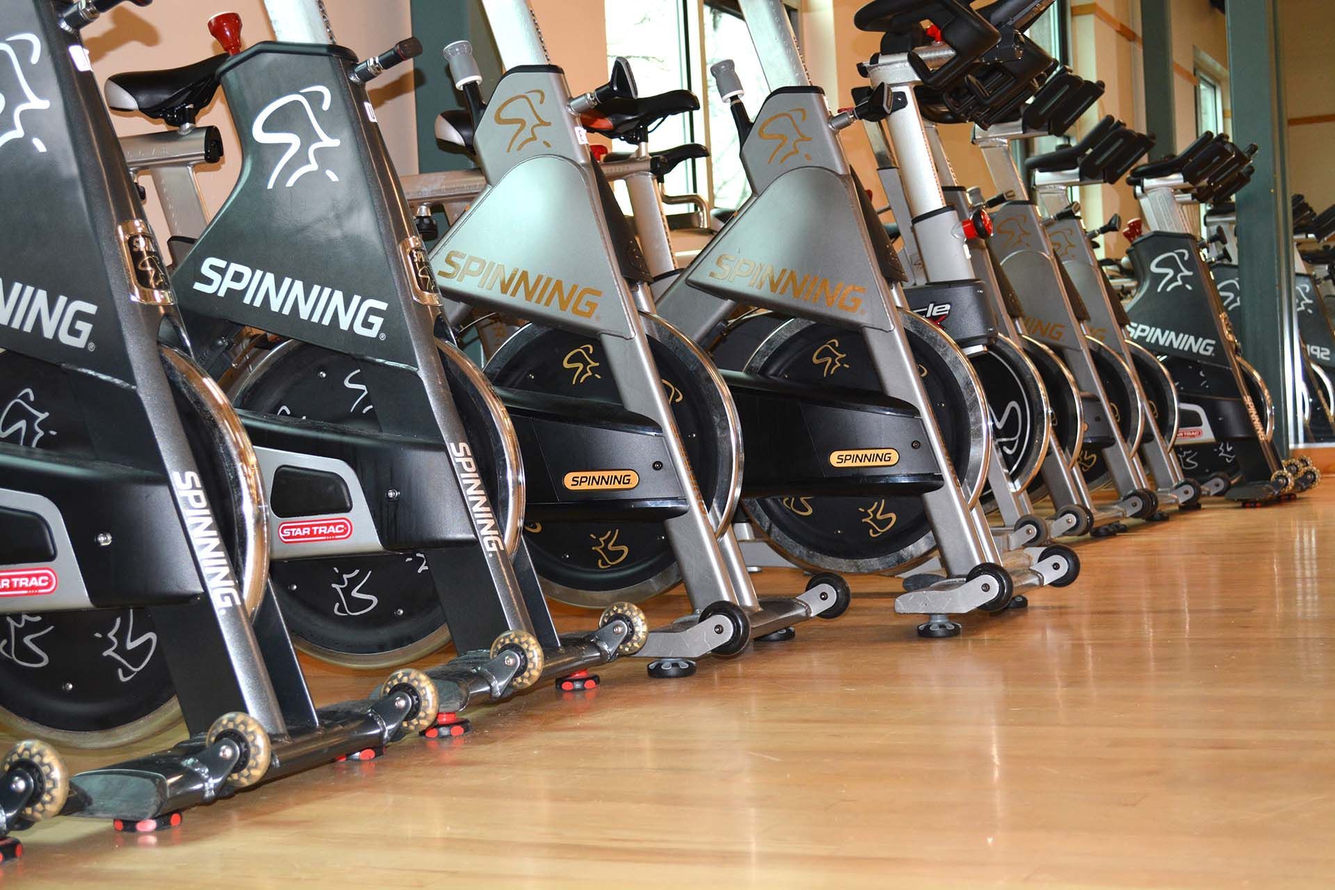 A row of black and silver indoor stationary exercise bikes lined up on a light wood floor in a fitness studio.