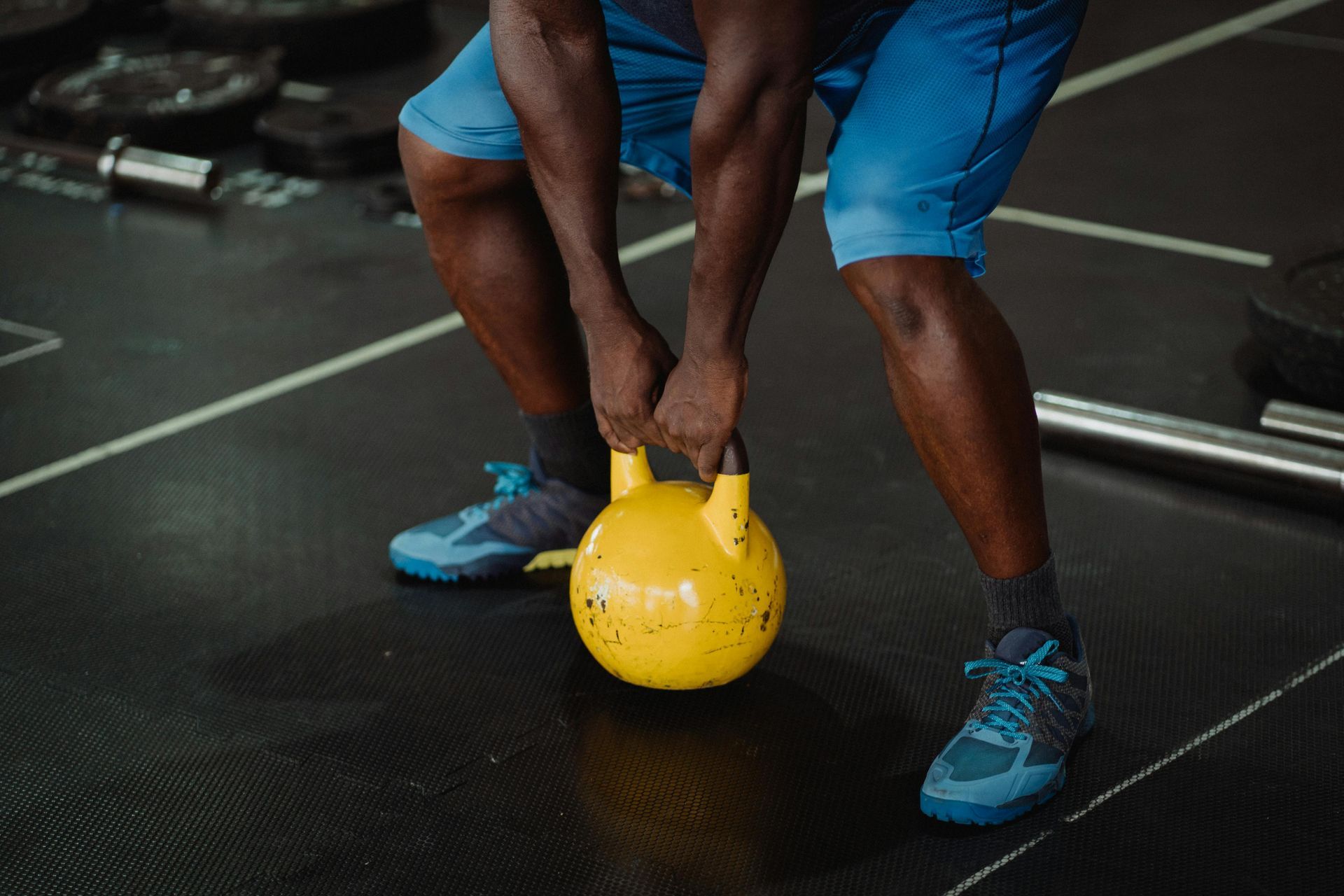 Man in blue shorts and shoes gripping yellow kettlebell, ready to lift in a gym.