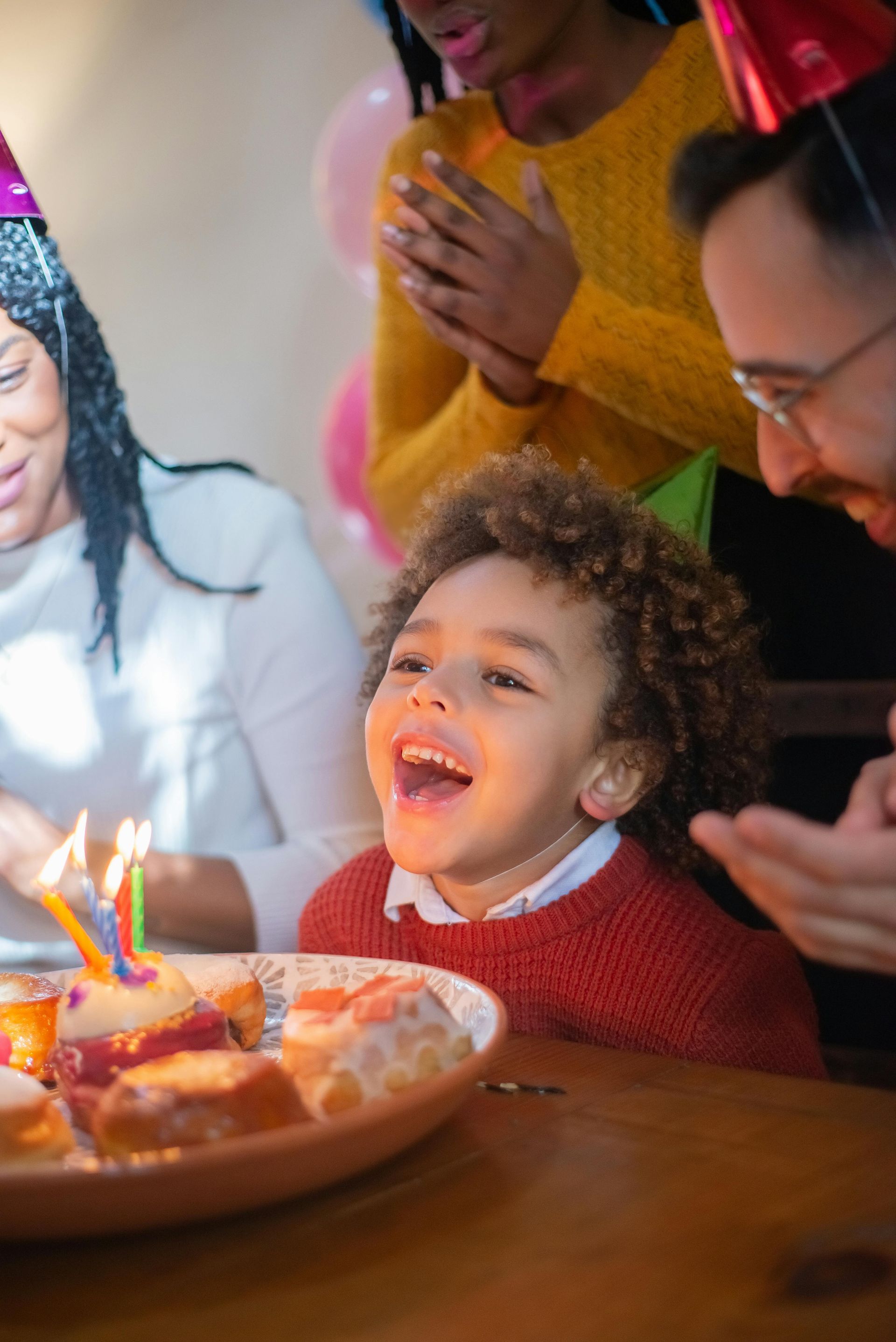 Child smiles at a birthday cake with lit candles; people clap and celebrate.