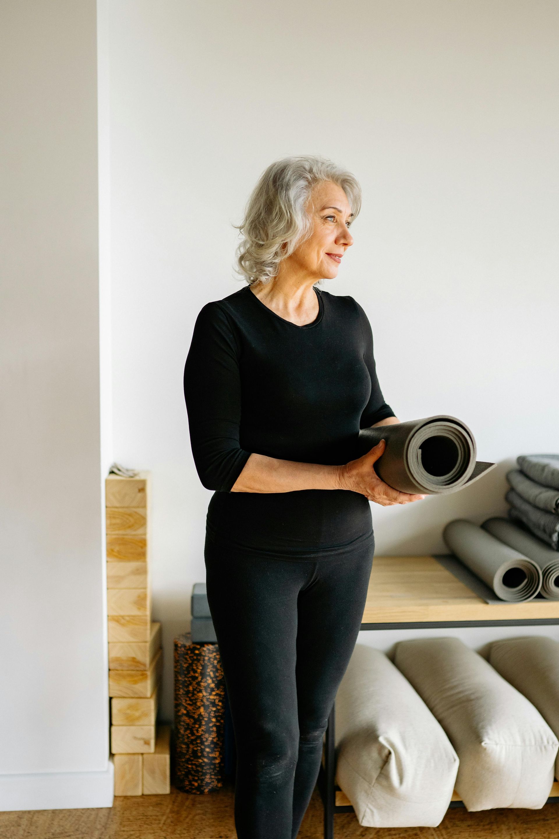 Woman in black workout clothes holding a rolled-up yoga mat, smiling, near mats and blocks.