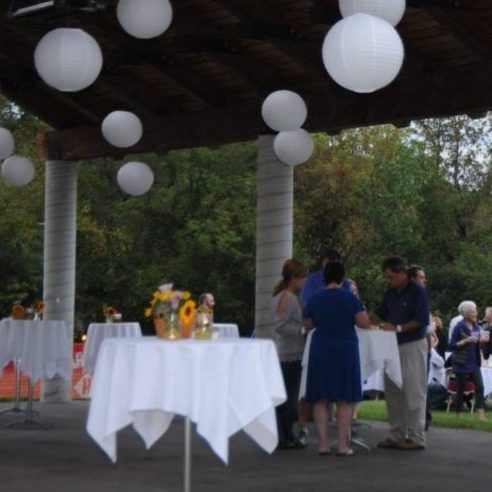 Outdoor event with white lanterns, tables, and people socializing under a wooden structure.