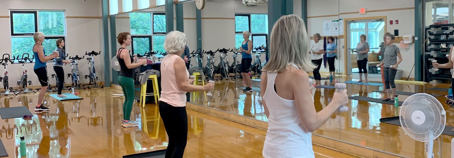 People exercising in a gym; some are holding small weights. A fan is visible in the foreground.