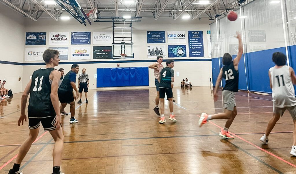 Basketball game in an indoor gym. Player in black shooting a ball toward the basket while other players look on.
