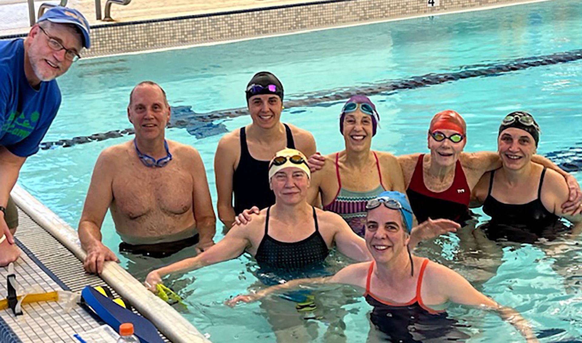 Group of swimmers smiling in a pool, posing near the edge. Some wearing swim caps and goggles.