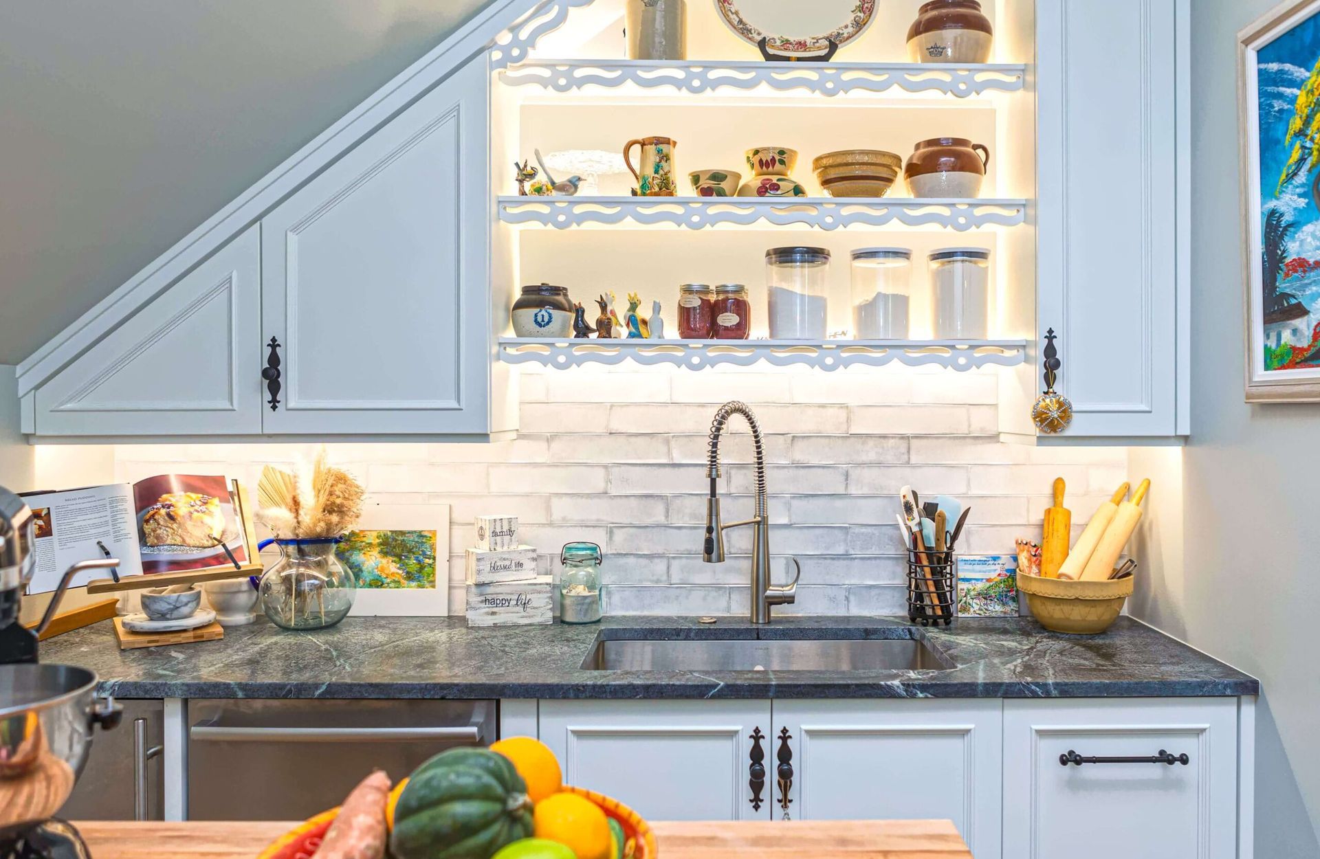 Kitchen with white cabinets, stone countertop, sink, and shelves with dishware and jars.