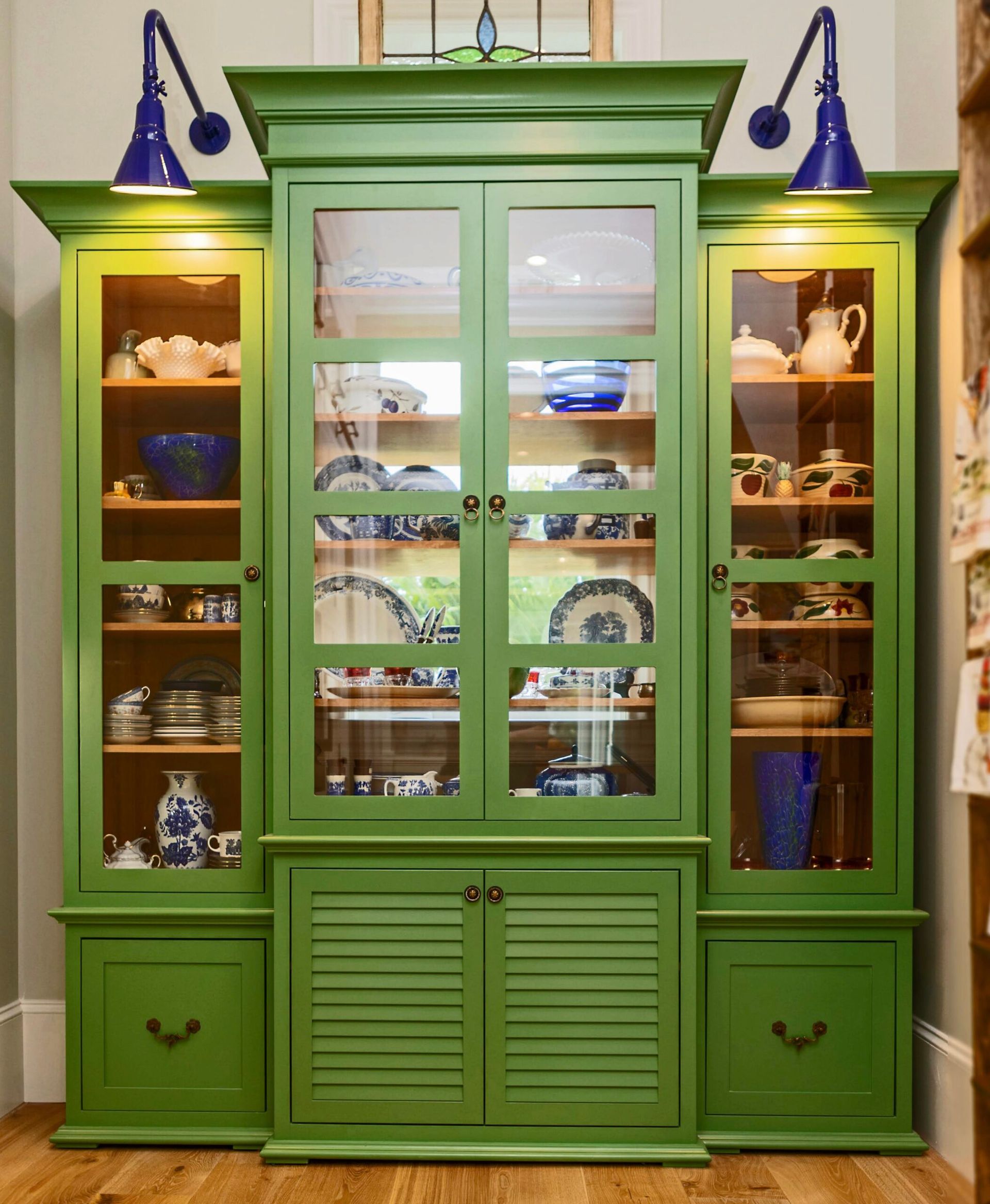 Green cabinet with glass doors displaying dishware, lit by blue wall sconces.