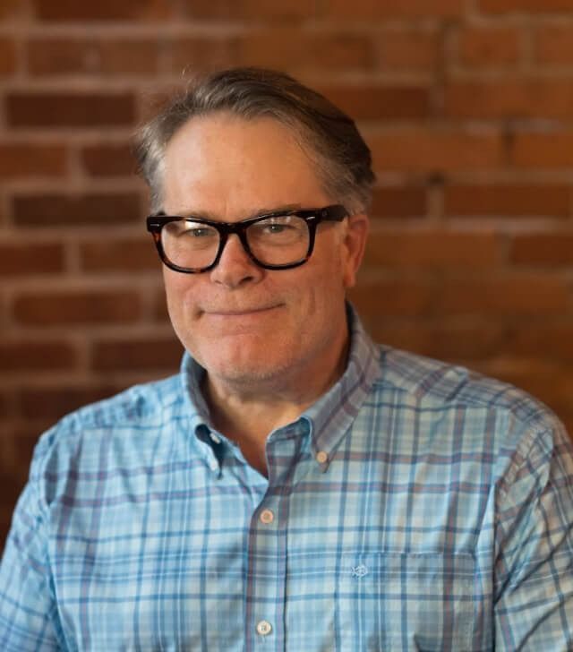 Man with glasses in blue plaid shirt, smiles in front of a brick wall.