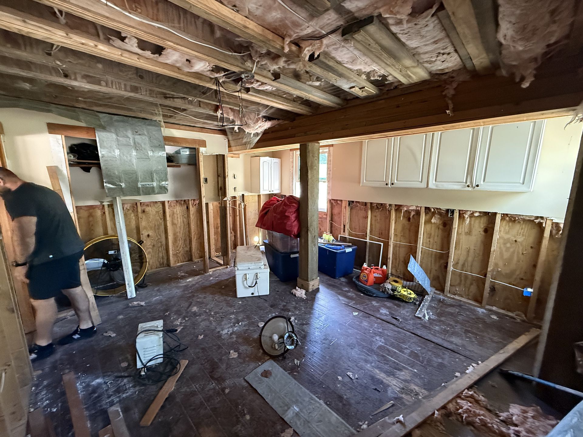 Interior view of a room under renovation. Walls stripped to studs, exposed ceiling beams, person working on the left.