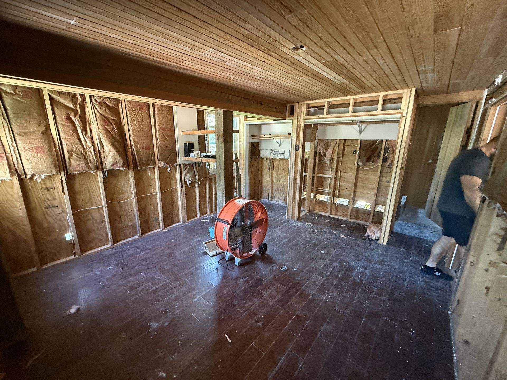 Room under renovation with exposed studs, a large fan, and a person near a window.
