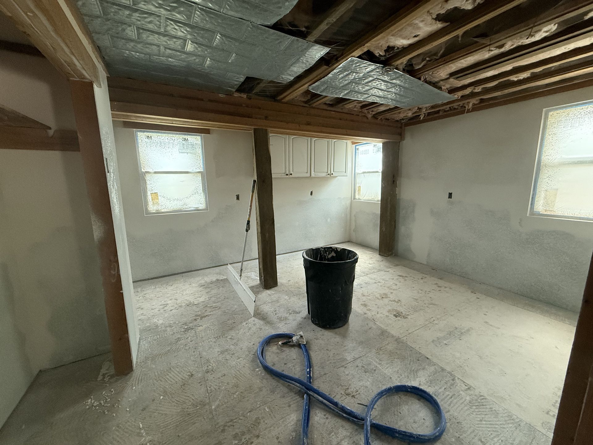Interior of a room under construction with bare walls, exposed beams, windows, and a black trash can.