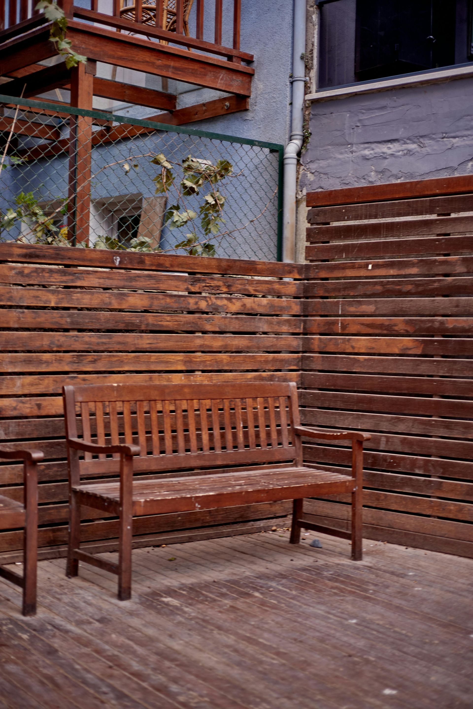 Wooden bench on a deck, surrounded by wood and fence. Blue building and balcony in the background.