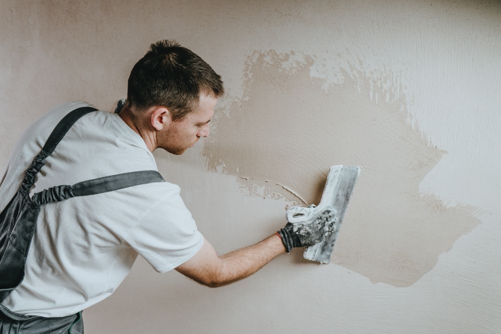 Man wearing overalls, applying plaster to a wall with a trowel.