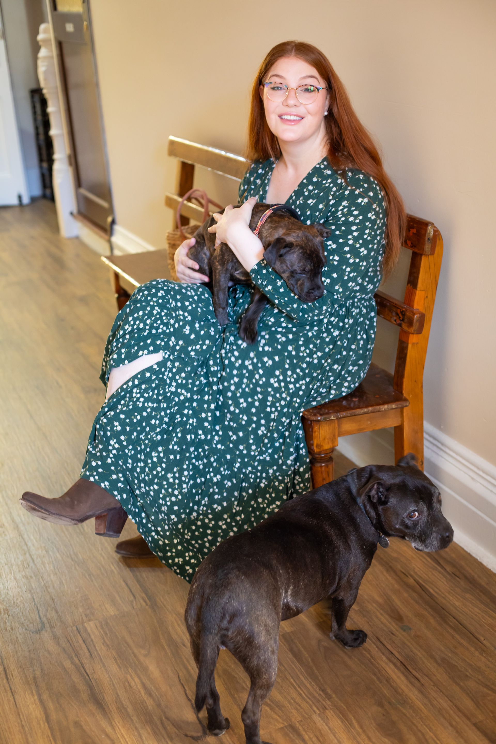 Dr Hannah Burn sitting on a wooden bench, holding a small dark dog, Happy; and Huxley stands nearby. She wears a green dress.
