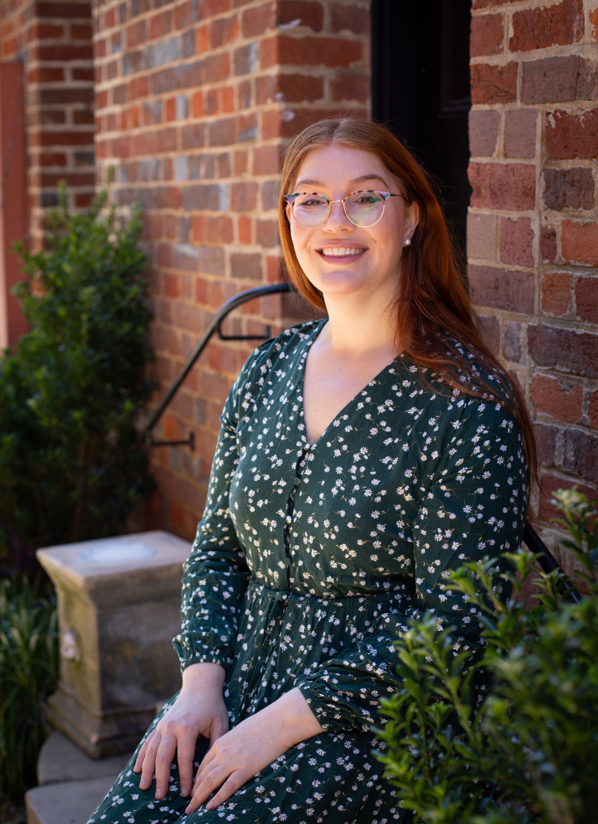 Dr Hannah Burn with red hair and glasses smiles in a green dress, sitting by a brick wall and bushes.