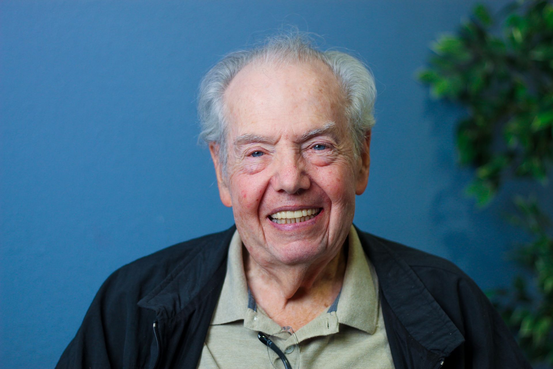Smiling older person wearing a light shirt and black jacket. They are set against a blue wall with a plant visible.