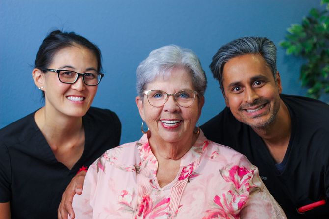 Two healthcare providers flank an older woman smiling, blue wall in background.