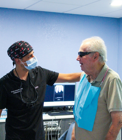Dentist with face mask comforting patient wearing bib and sunglasses. Dental office, x-ray on screen.