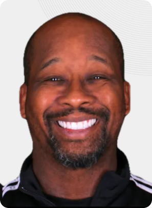 Man smiling, close-up, black track jacket, graying beard, white teeth, neutral background.