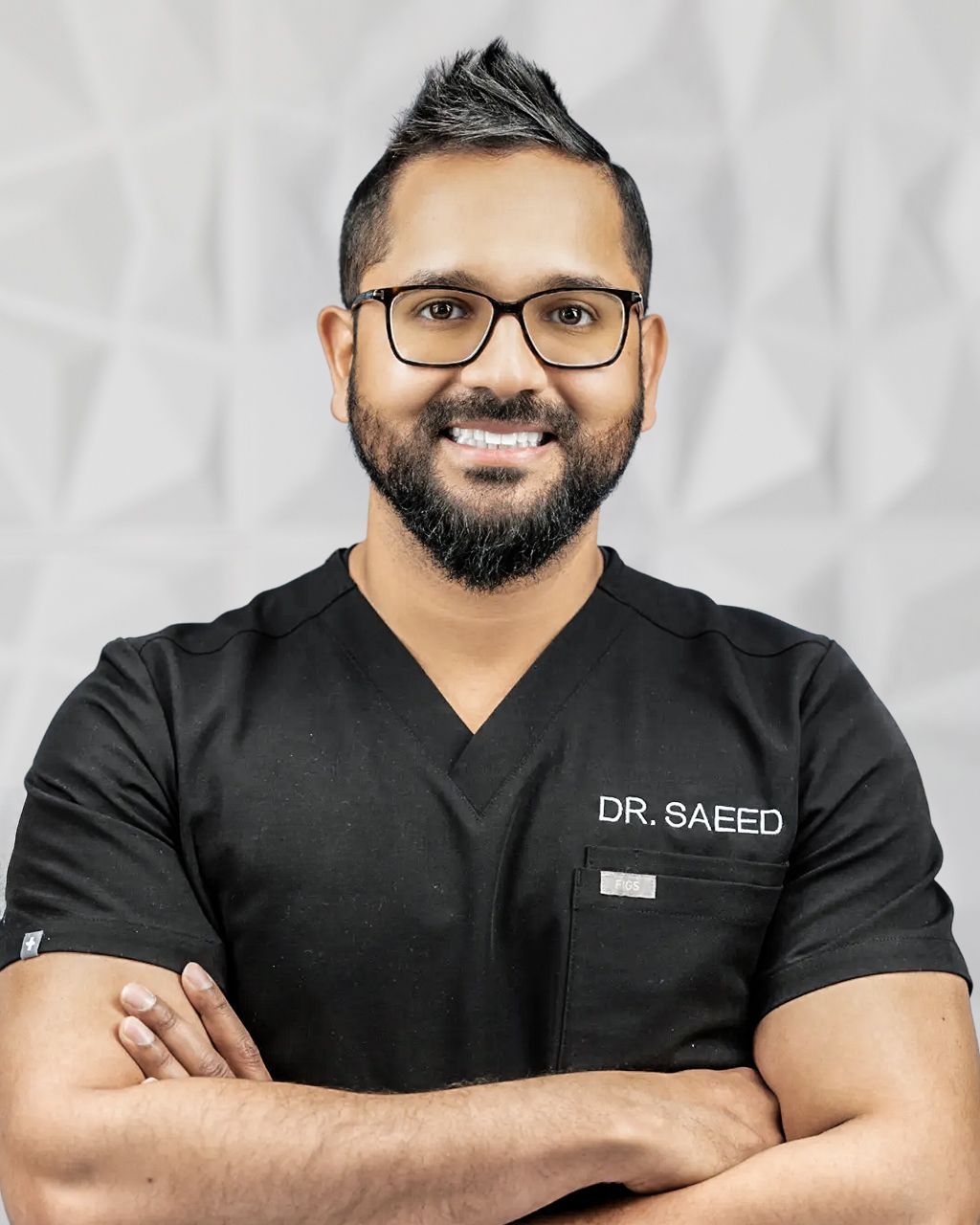 Smiling doctor in black scrubs with crossed arms, wearing glasses, in front of textured white wall.