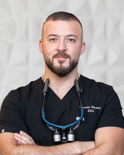 Man in black scrubs with crossed arms, wearing dental loupes, in a modern, white-walled setting.