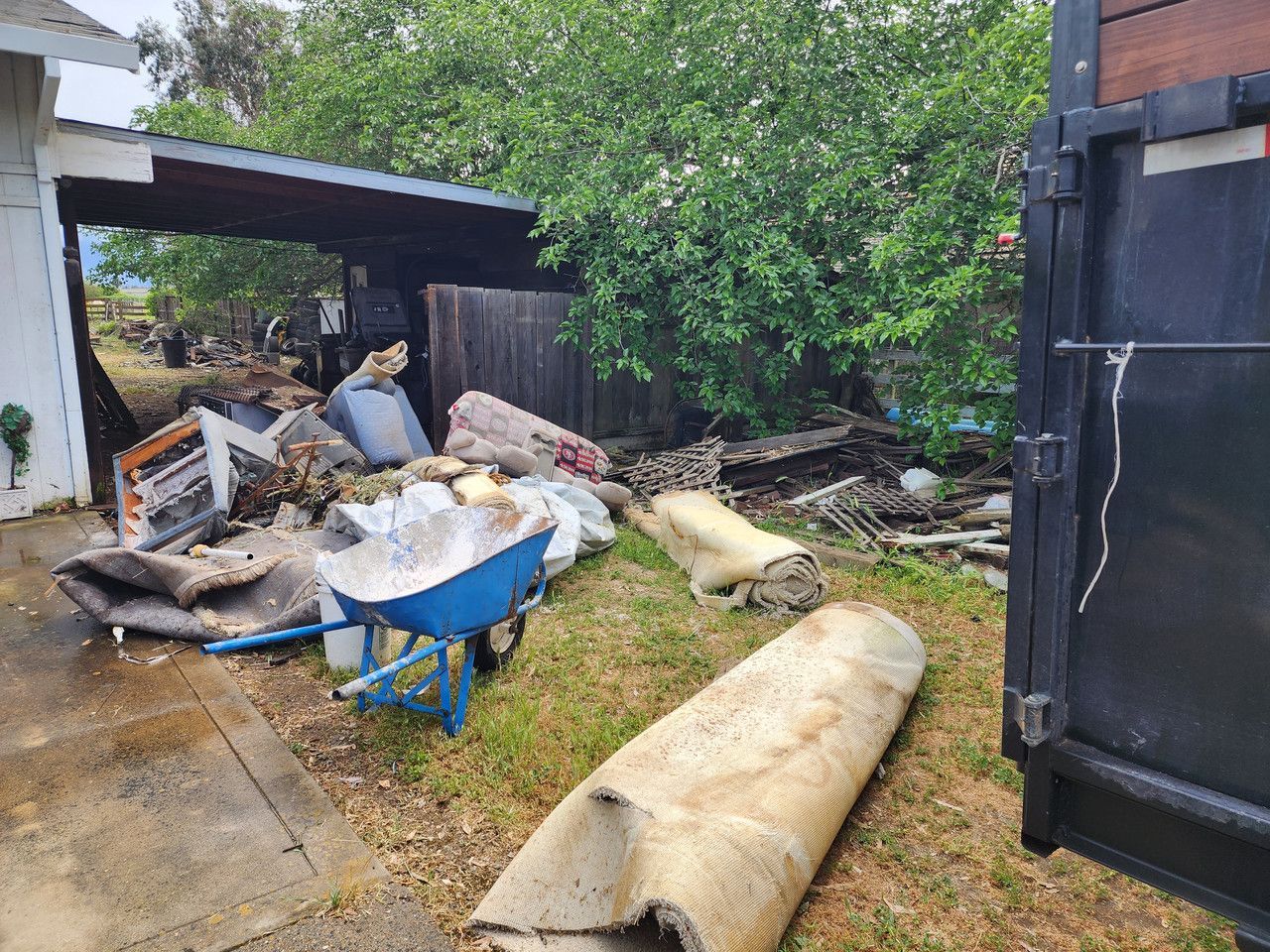pile of trash on side of house in the grass. Trash items include carpets, chairs, a wheelbarrow and various other items