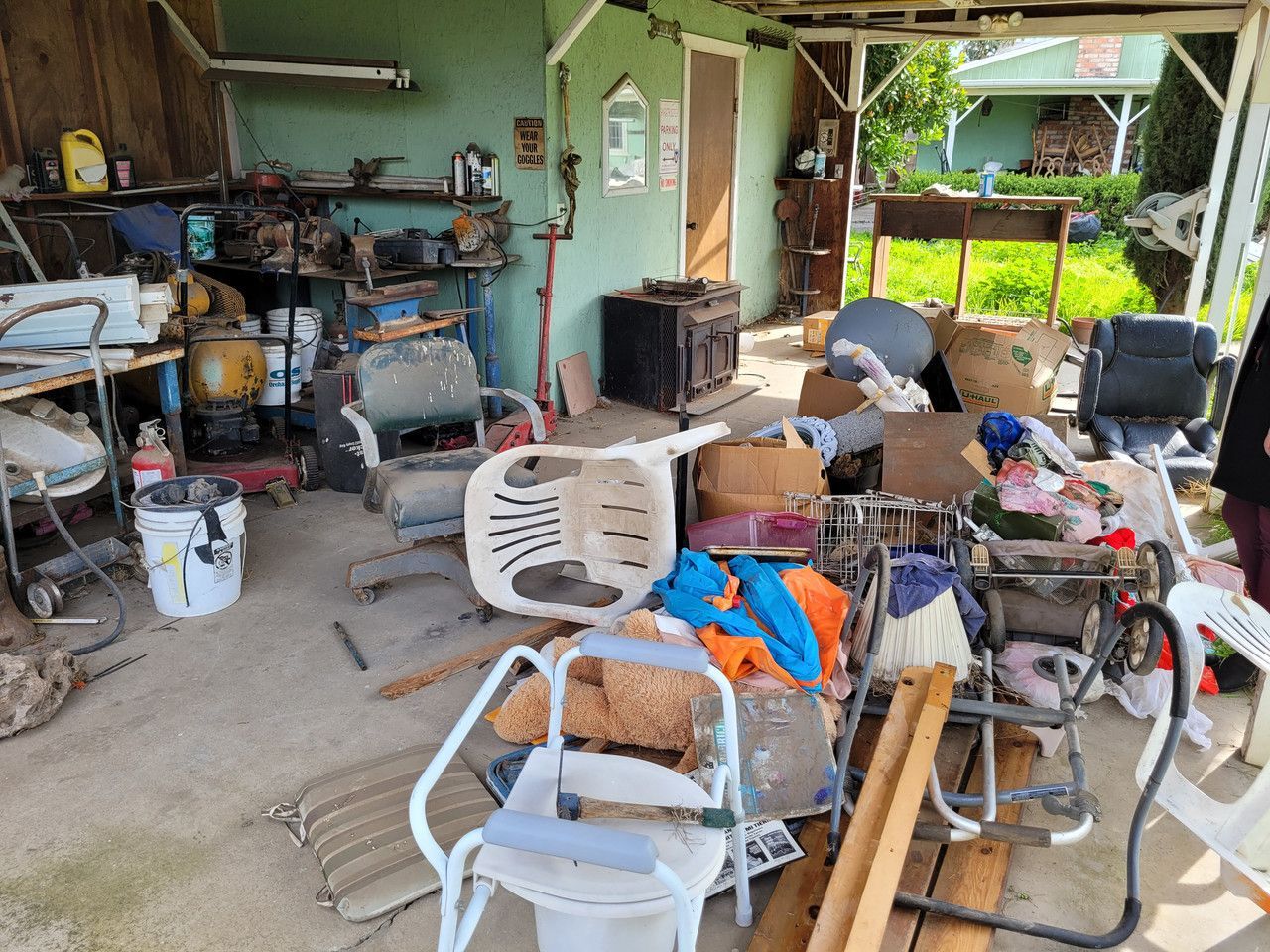 Backyard filled with a chaotic pile of discarded items including lawn chairs, a bed pan, worn-out clothing, stuffed animals, rusty tools, and broken lawn equipment, showcasing the aftermath of an eviction.