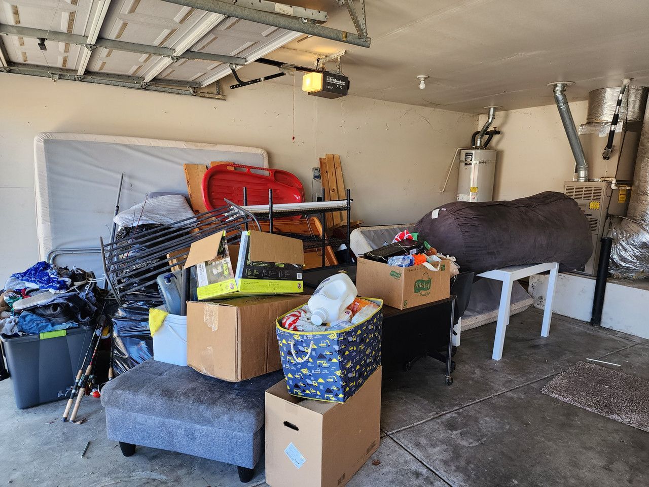 Garage filled with an assortment of household items including mattresses, fishing poles, stacked boxes, and miscellaneous clutter, representing a space in need of cleanout.