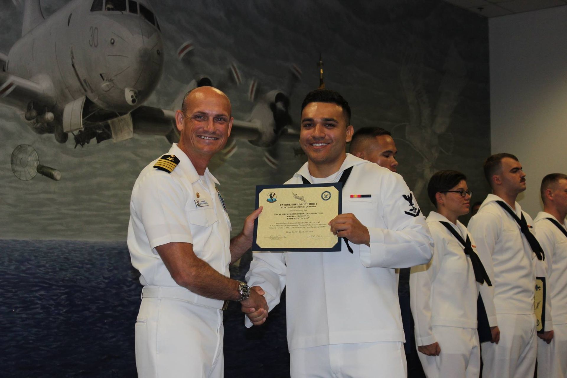 A man in a white uniform is shaking hands with a man in a navy uniform holding a certificate.