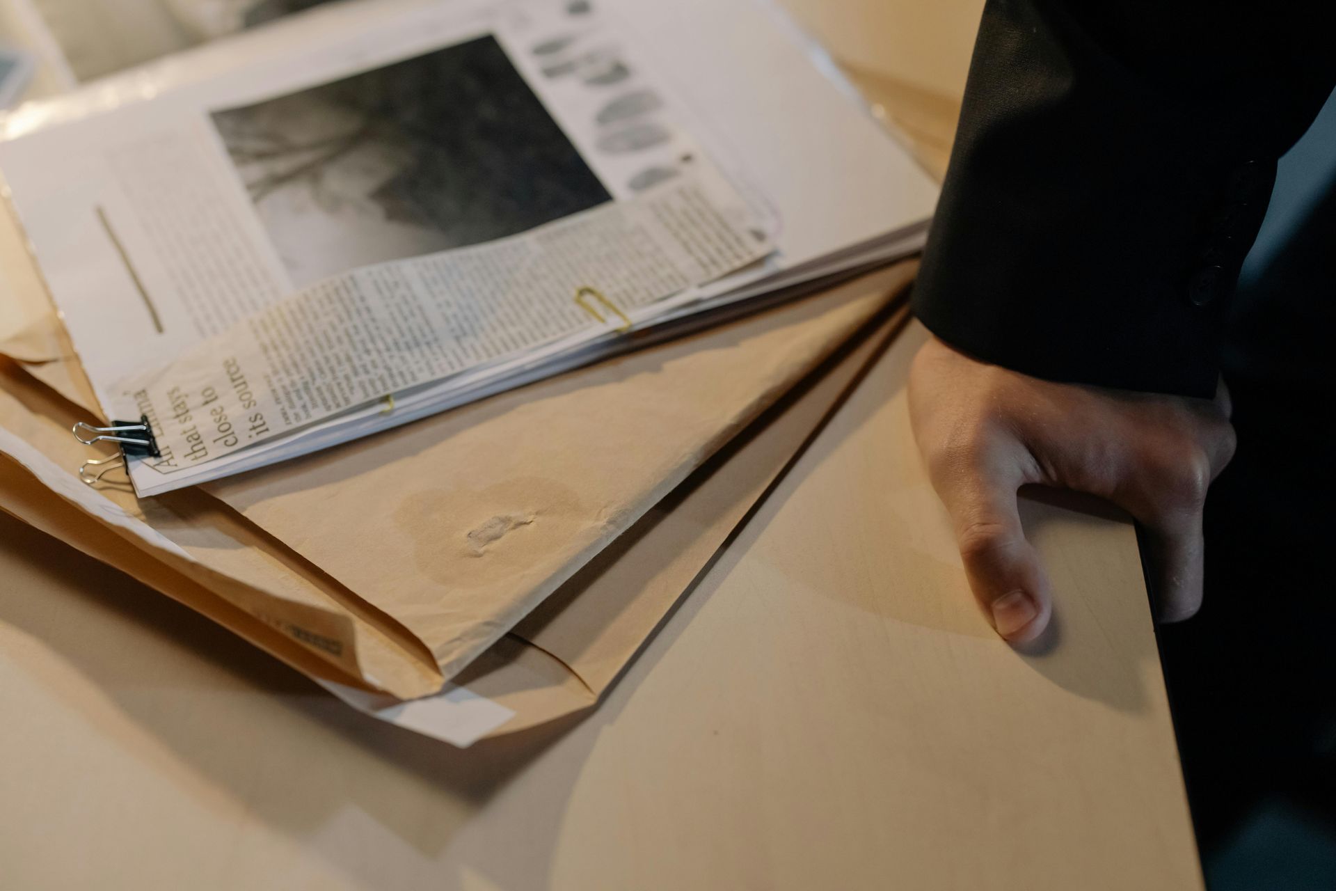 Person's hand on a stack of tan file folders, papers, and newsprint on a table.