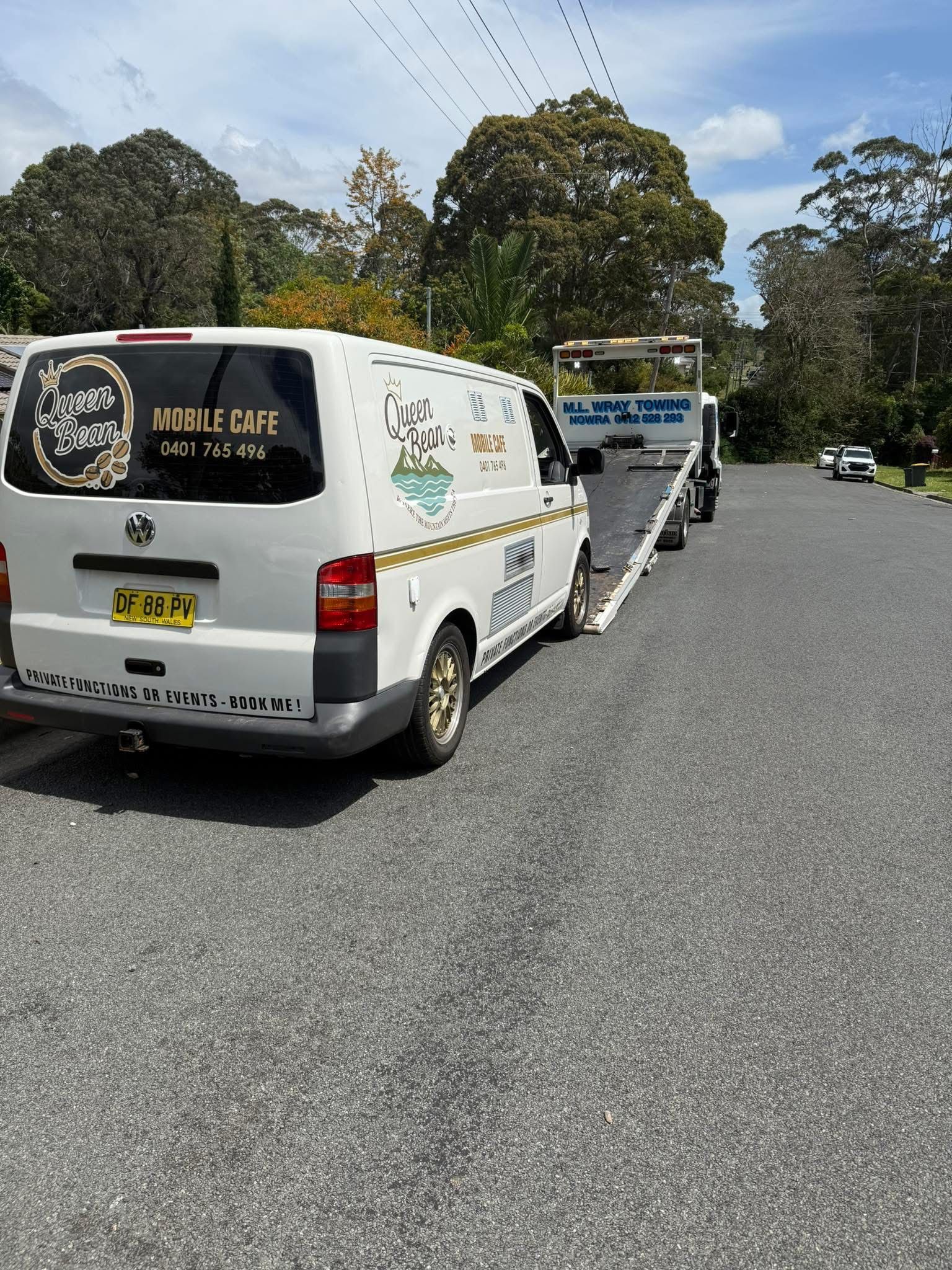 White van towing a flatbed trailer on a paved road. Trees and blue sky are in the background. — M.L. Wray Towing & Automotive Repairs in Nowra, NSW